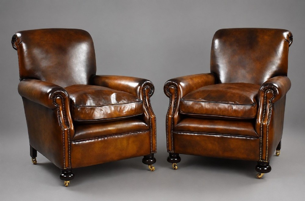 A pair of antique chestnut brown leather club chairs featuring rolled arms, brass stud trimming, loose cushion seats, and dark turned legs on brass castors.