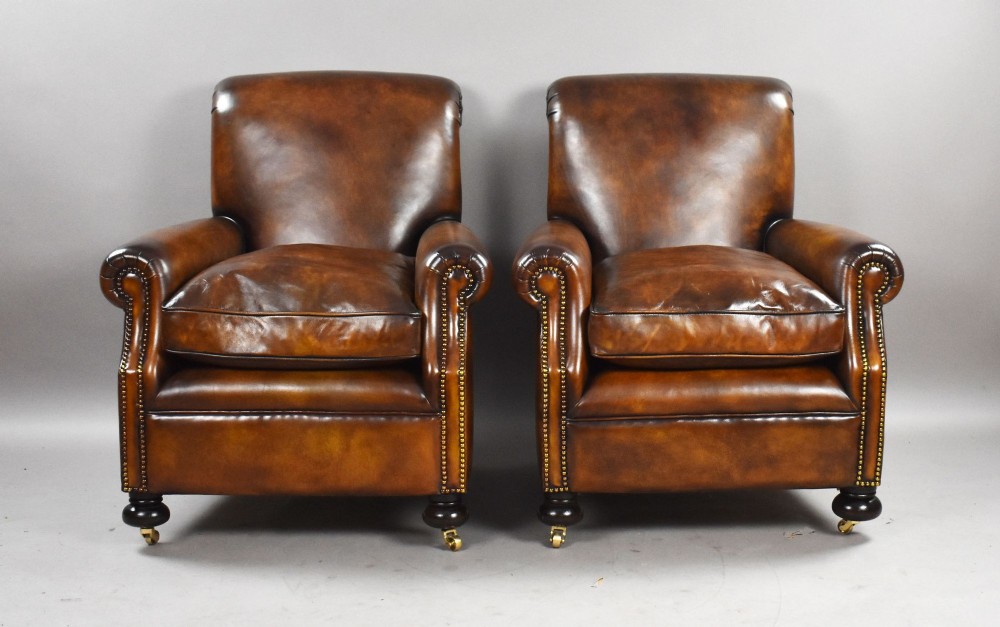 A pair of antique chestnut brown leather club chairs featuring rolled arms, brass stud trimming, loose cushion seats, and dark turned legs on brass castors. View II