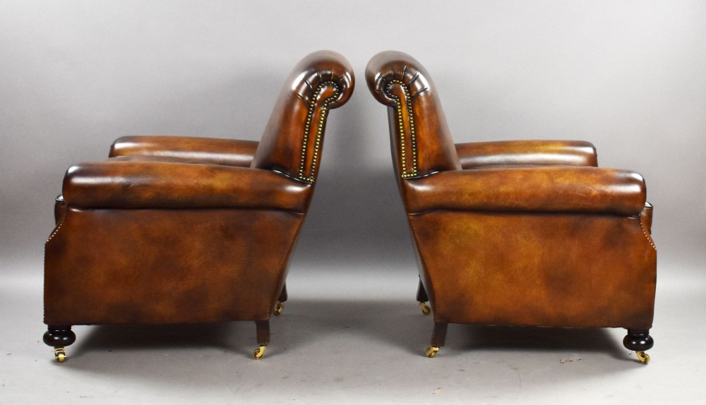 A pair of antique chestnut brown leather club chairs featuring rolled arms, brass stud trimming, loose cushion seats, and dark turned legs on brass castors. View III