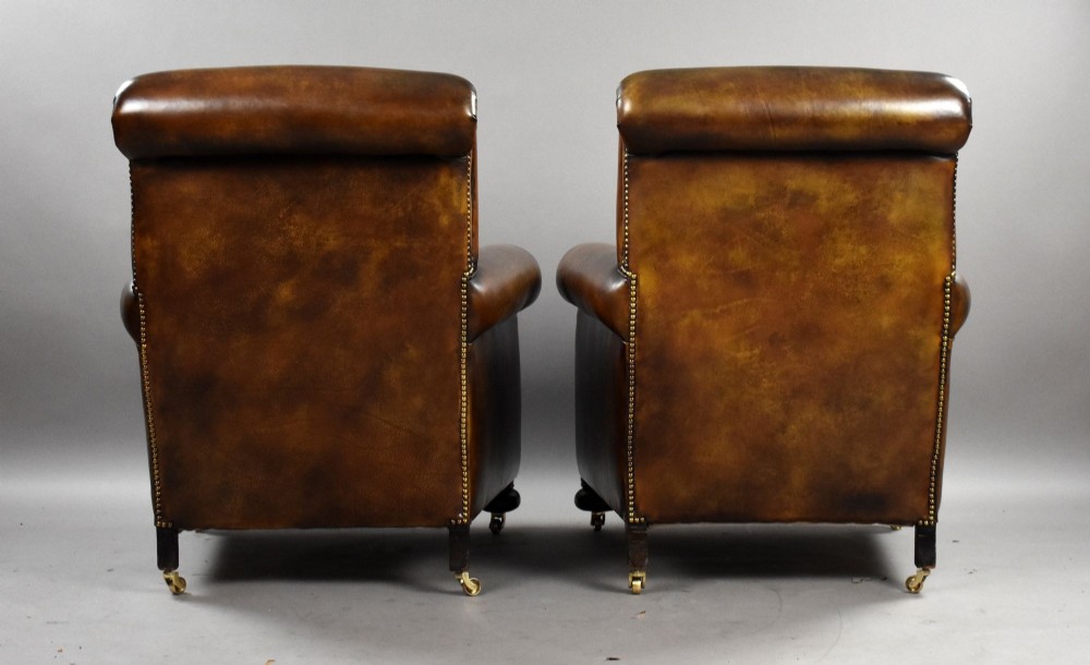 A pair of antique chestnut brown leather club chairs featuring rolled arms, brass stud trimming, loose cushion seats, and dark turned legs on brass castors. View IV