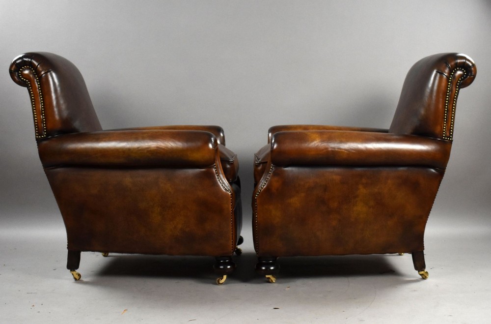 A pair of antique chestnut brown leather club chairs featuring rolled arms, brass stud trimming, loose cushion seats, and dark turned legs on brass castors. View V