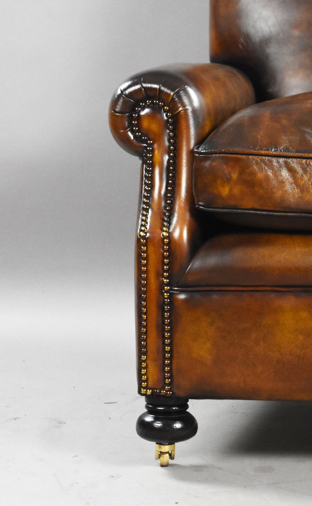 A pair of antique chestnut brown leather club chairs featuring rolled arms, brass stud trimming, loose cushion seats, and dark turned legs on brass castors. View VIII