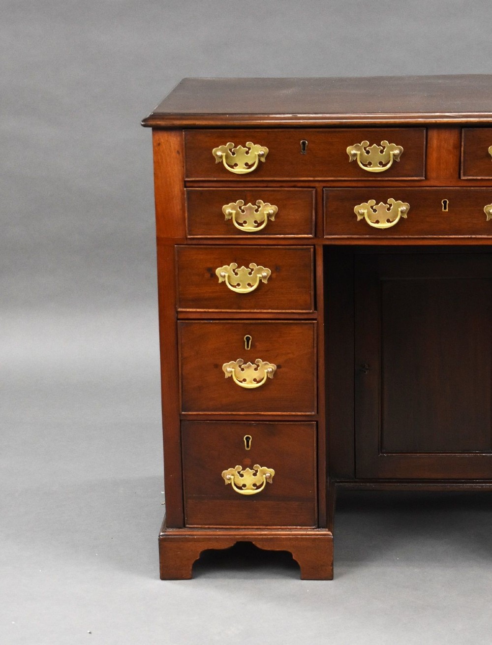 A George III mahogany kneehole desk featuring a flat rectangular top, brass swan-neck handles on multiple drawers, a central recessed cupboard, and bracket feet. View III