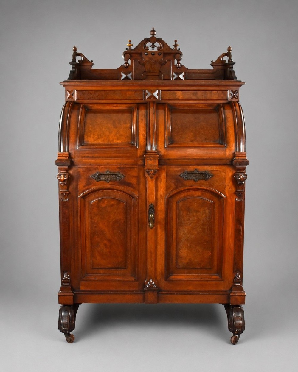 A Victorian walnut Wooton secretary desk featuring carved cresting, curved upper doors, panelling, brass mail slots, and carved feet on castors.