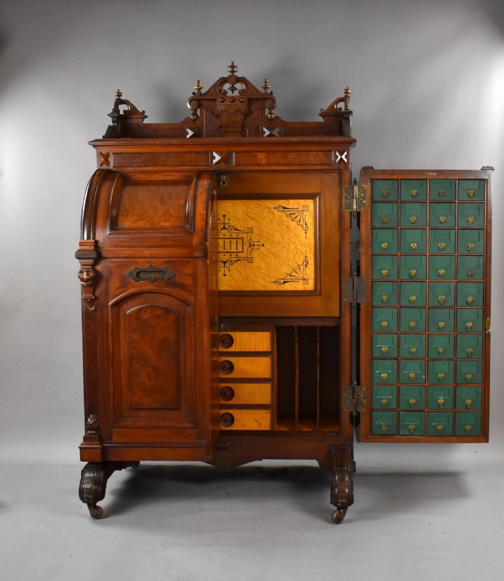 A Victorian walnut Wooton secretary desk featuring carved cresting, curved upper doors, panelling, brass mail slots, and carved feet on castors. View II