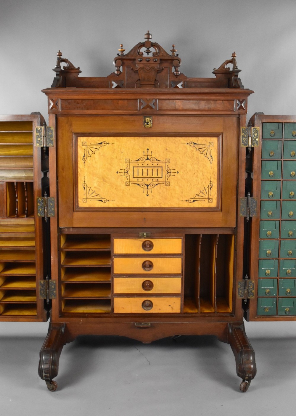 A Victorian walnut Wooton secretary desk featuring carved cresting, curved upper doors, panelling, brass mail slots, and carved feet on castors. View VI