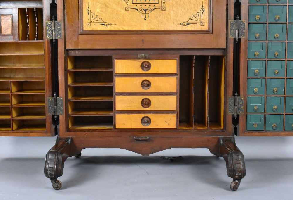 A Victorian walnut Wooton secretary desk featuring carved cresting, curved upper doors, panelling, brass mail slots, and carved feet on castors. View VIII