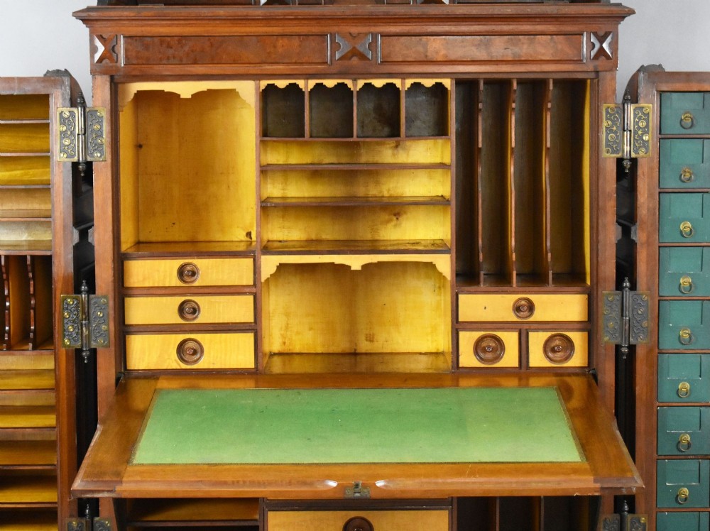 A Victorian walnut Wooton secretary desk featuring carved cresting, curved upper doors, panelling, brass mail slots, and carved feet on castors. View X