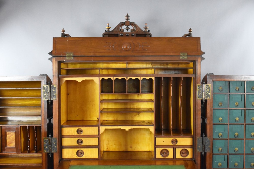 A Victorian walnut Wooton secretary desk featuring carved cresting, curved upper doors, panelling, brass mail slots, and carved feet on castors. View XI