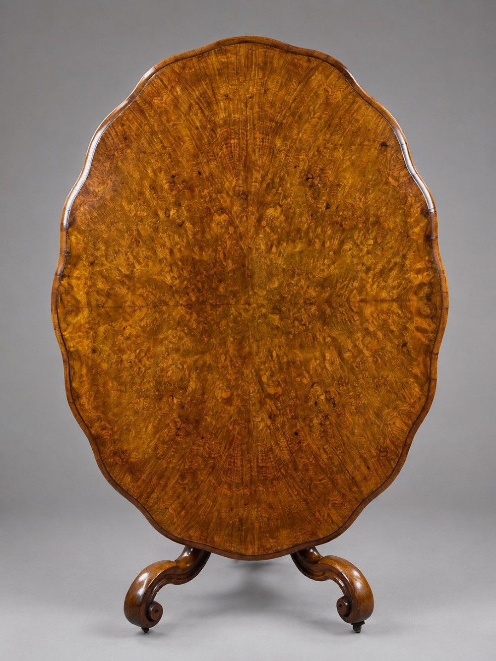 A Victorian tilt-top loo table shown vertically, featuring quarter-matched burr walnut veneer with a scalloped edge on carved cabriole legs.