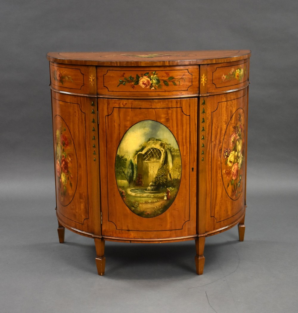 An Edwardian demi-lune satinwood cabinet featuring hand-painted floral and landscape medallions on two doors and tapered legs. View II