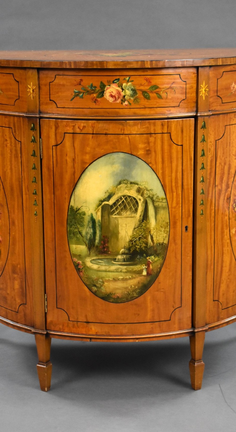 An Edwardian demi-lune satinwood cabinet featuring hand-painted floral and landscape medallions on two doors and tapered legs. View IV