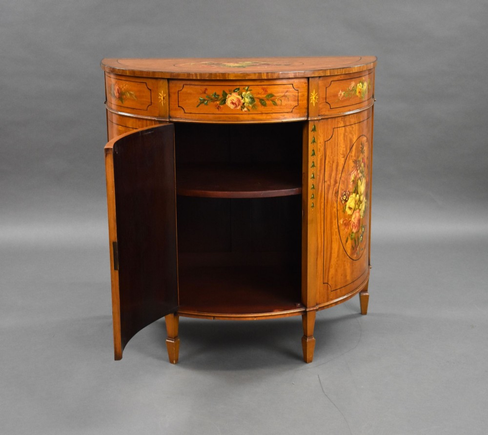 An Edwardian demi-lune satinwood cabinet featuring hand-painted floral and landscape medallions on two doors and tapered legs. View VI