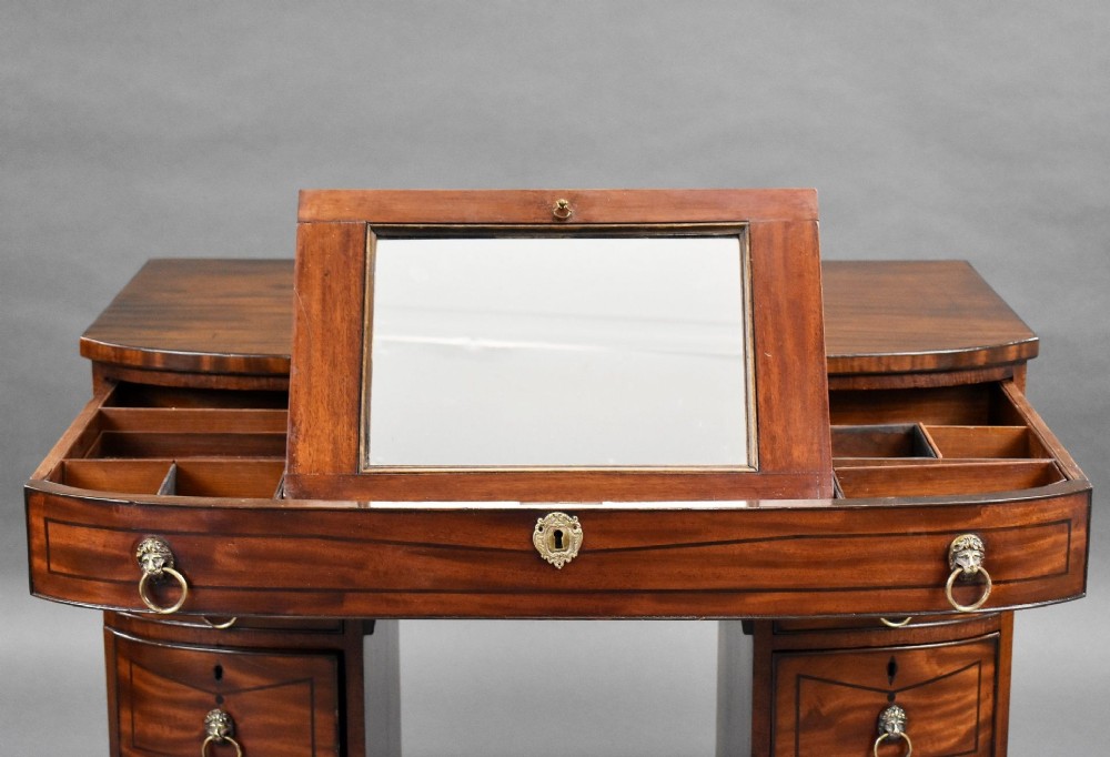 An antique Regency flame mahogany kneehole dressing chest with seven drawers, brass lion mask ring handles, and turned feet. View X