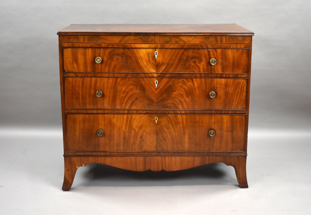 A Regency mahogany chest of drawers featuring three full-length drawers with brass knobs, bone keyhole escutcheons, flame veneer fronts, and a shaped apron base. View II