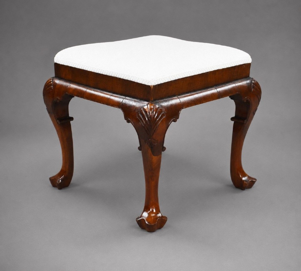 A square Queen Anne style walnut stool featuring cream textured fabric upholstery and carved cabriole legs with shell motifs.
