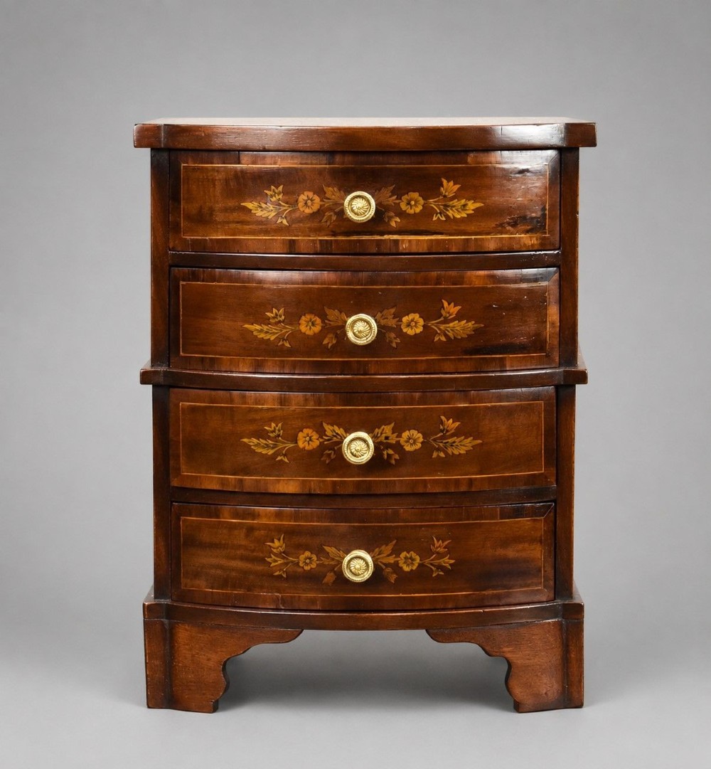 A miniature warm brown wooden chest of drawers featuring a bow front, four drawers with floral marquetry inlay, and central brass knobs.