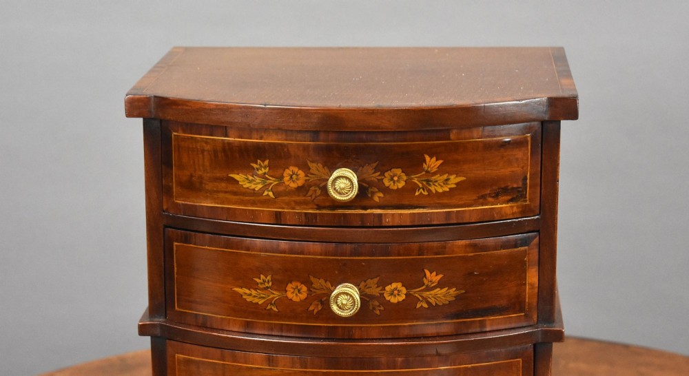 A miniature warm brown wooden chest of drawers featuring a bow front, four drawers with floral marquetry inlay, and central brass knobs. View III