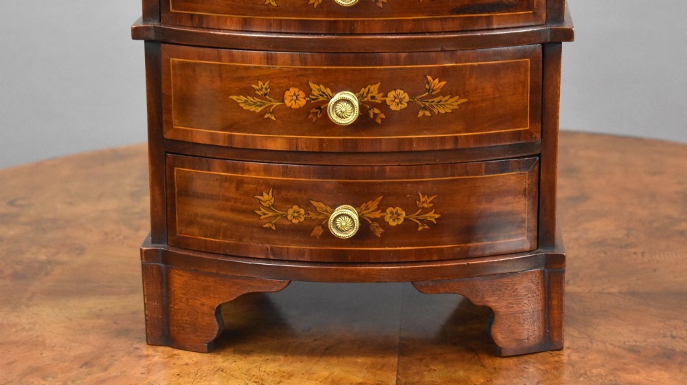 A miniature warm brown wooden chest of drawers featuring a bow front, four drawers with floral marquetry inlay, and central brass knobs. View IV