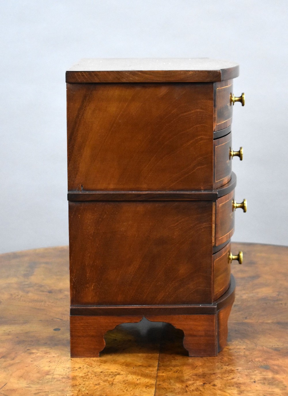 A miniature warm brown wooden chest of drawers featuring a bow front, four drawers with floral marquetry inlay, and central brass knobs. View V
