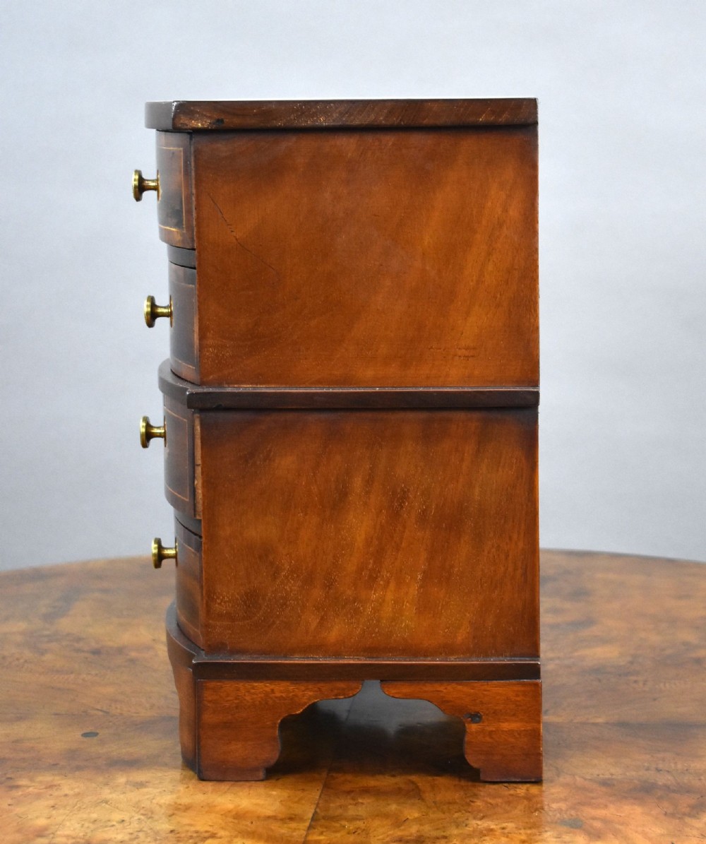A miniature warm brown wooden chest of drawers featuring a bow front, four drawers with floral marquetry inlay, and central brass knobs. View VI