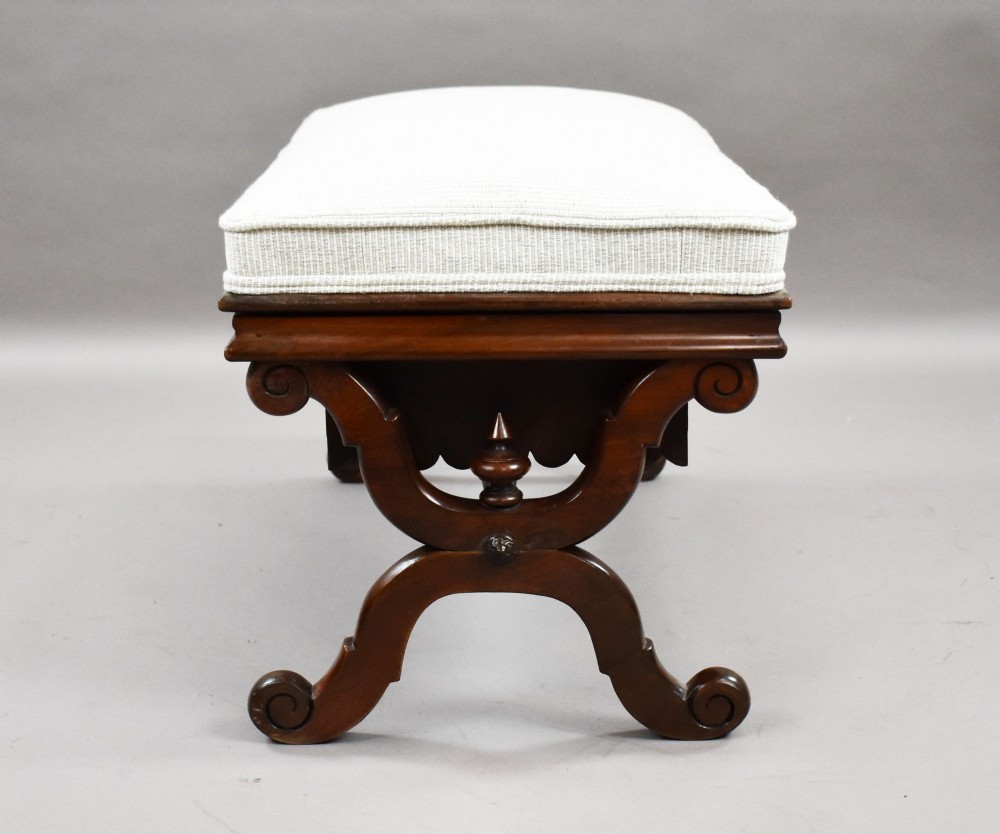 A William IV mahogany stool featuring a scalloped apron, carved scroll end supports, a turned central stretcher, and an off-white upholstered seat cushion. View V