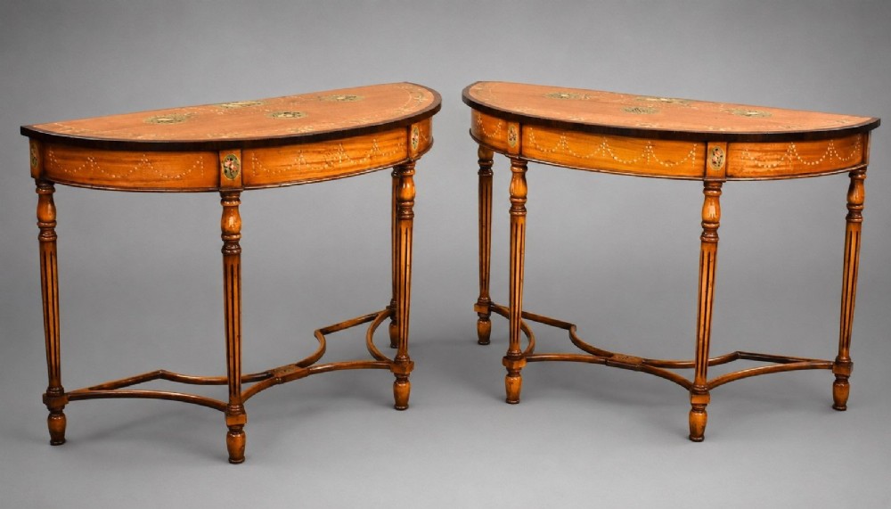 A pair of Sheraton Revival demi-lune console tables in golden satinwood with hand-painted floral garlands, fluted tapered legs, and decorative stretchers.