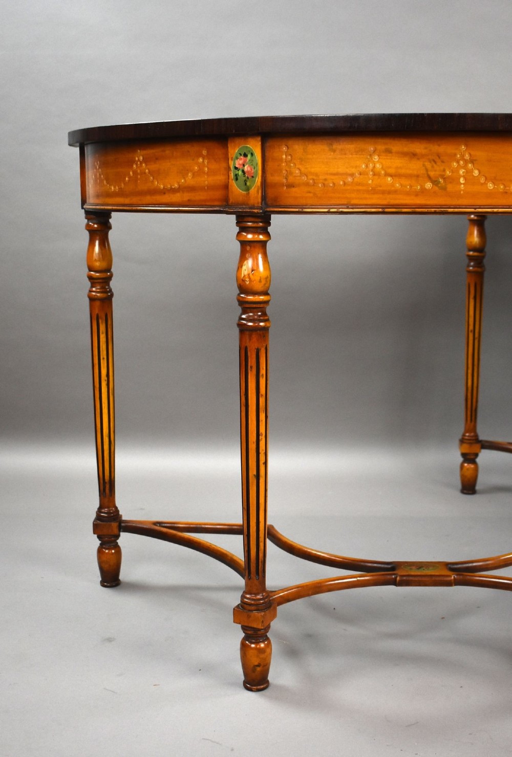 A pair of Sheraton Revival demi-lune console tables in golden satinwood with hand-painted floral garlands, fluted tapered legs, and decorative stretchers. View III