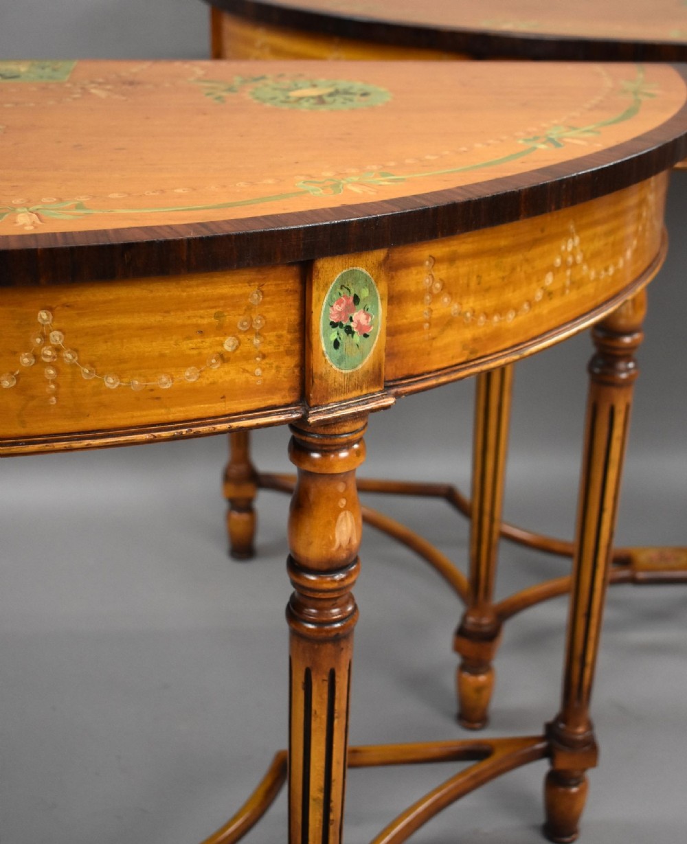 A pair of Sheraton Revival demi-lune console tables in golden satinwood with hand-painted floral garlands, fluted tapered legs, and decorative stretchers. View V