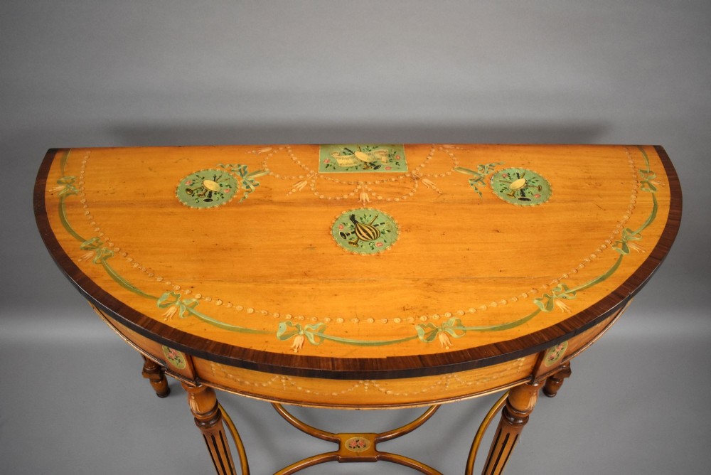 A pair of Sheraton Revival demi-lune console tables in golden satinwood with hand-painted floral garlands, fluted tapered legs, and decorative stretchers. View VI