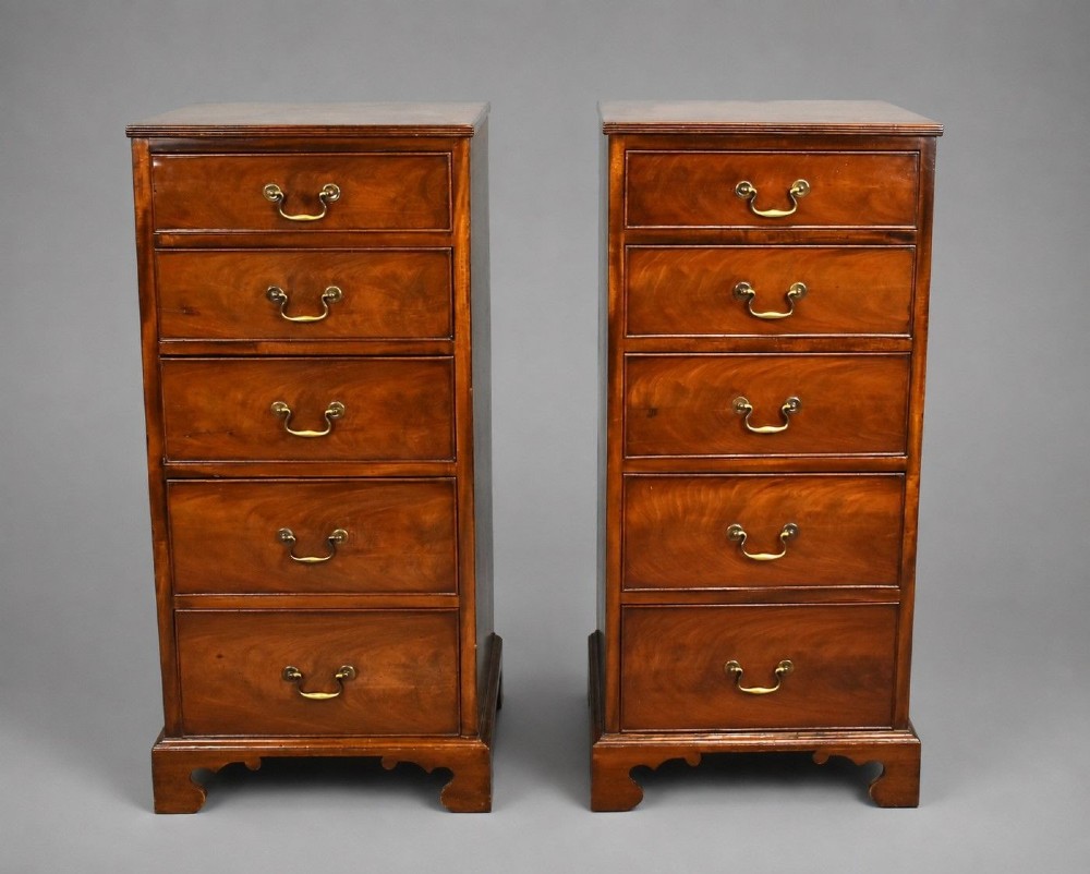 A matching pair of tall, narrow mahogany bedside chests, each featuring five drawers with brass handles and resting on bracket feet.