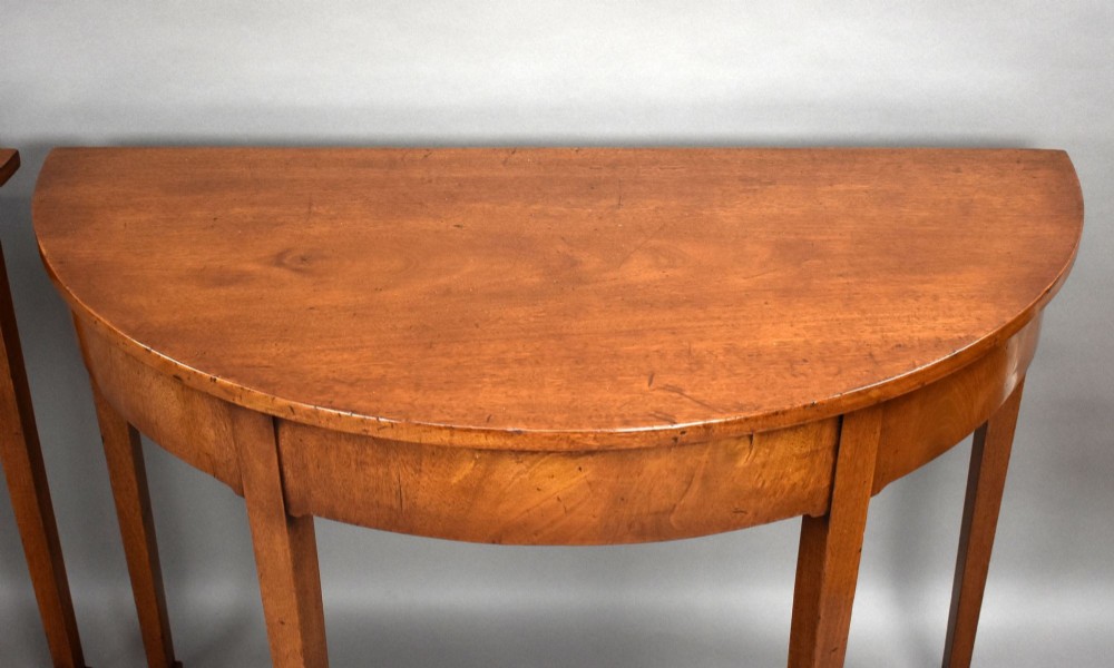 A pair of Georgian mahogany demilune console tables featuring semi-circular tops, plain aprons, and square tapered legs. View V