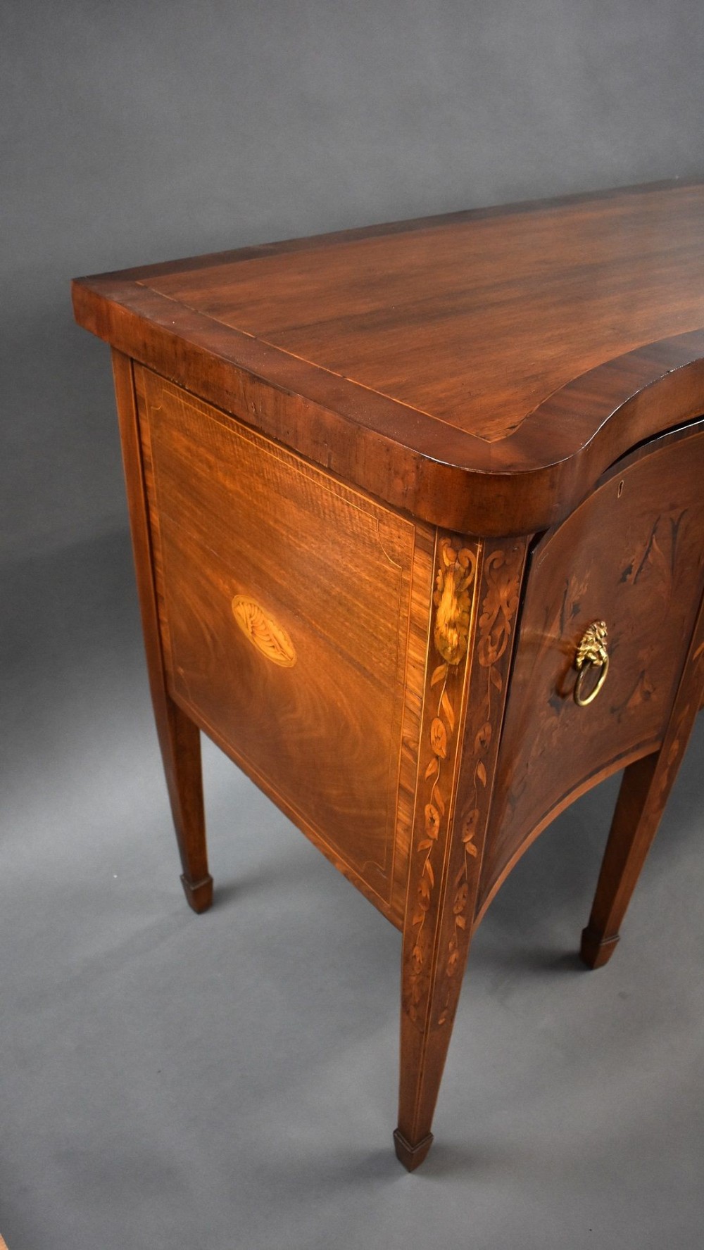 An antique mahogany serpentine sideboard featuring floral marquetry inlays, a central drawer, two side cupboards, tapered square legs, and brass lion-mask ring handles. View V
