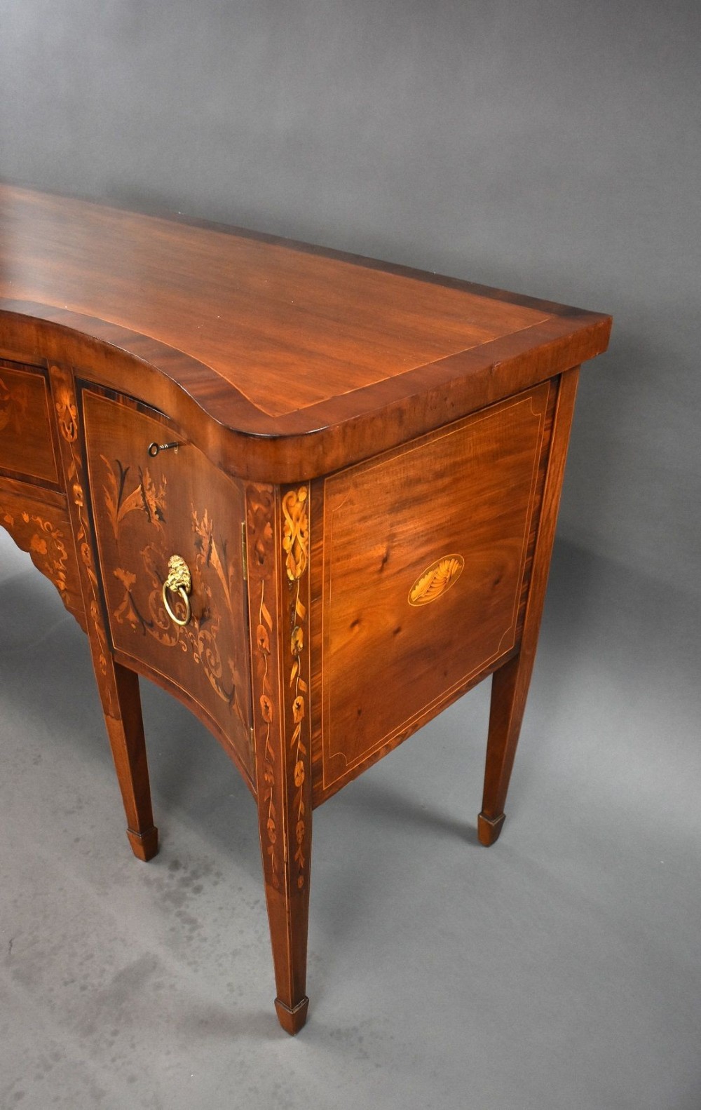 An antique mahogany serpentine sideboard featuring floral marquetry inlays, a central drawer, two side cupboards, tapered square legs, and brass lion-mask ring handles. View VI