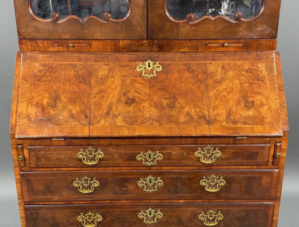 A George II burr walnut bureau bookcase featuring an upper double-door cabinet with shaped mirrored panels over a fall-front desk and four drawers with brass handles. View VI