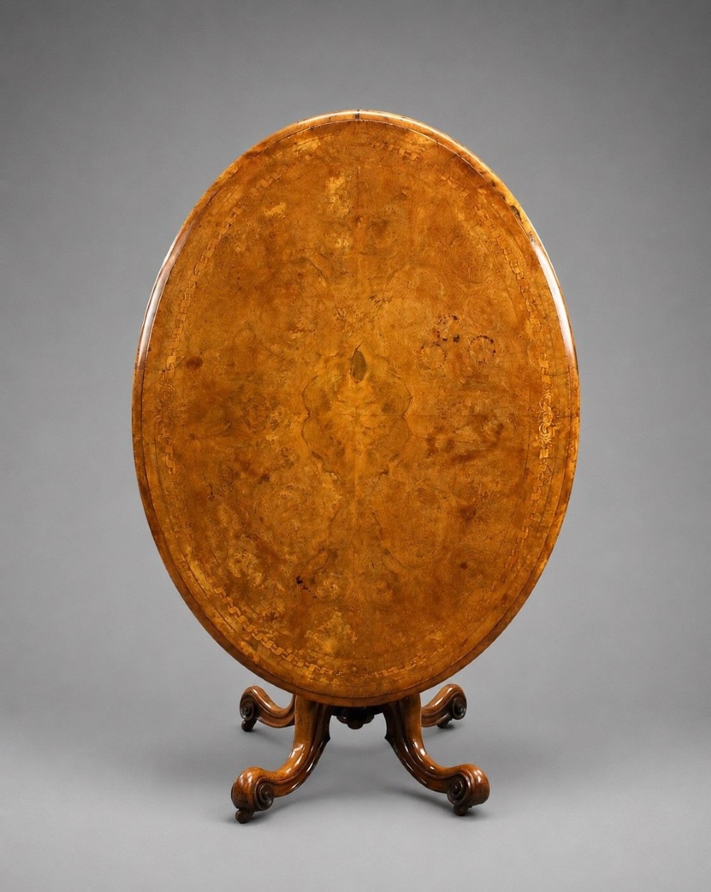 An oval Victorian burr walnut loo table featuring geometric and floral marquetry inlay, shown upright via a tilt-top mechanism on a carved four-legged pedestal base.