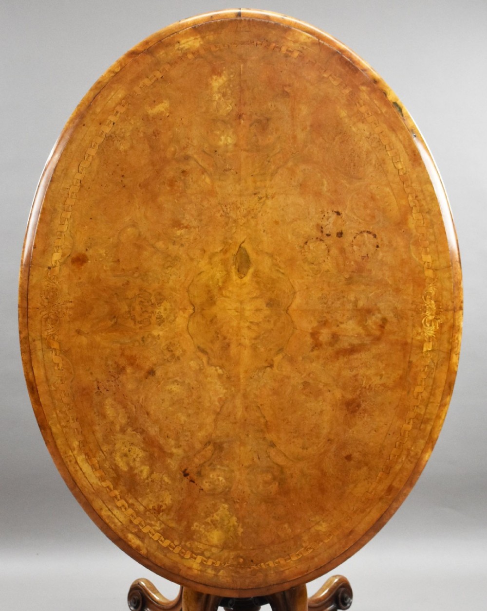 An oval Victorian burr walnut loo table featuring geometric and floral marquetry inlay, shown upright via a tilt-top mechanism on a carved four-legged pedestal base. View II