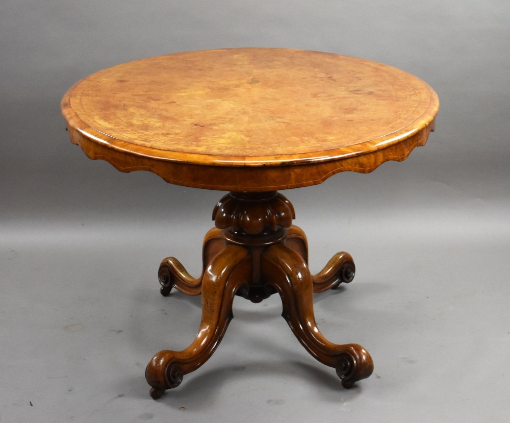 An oval Victorian burr walnut loo table featuring geometric and floral marquetry inlay, shown upright via a tilt-top mechanism on a carved four-legged pedestal base. View III