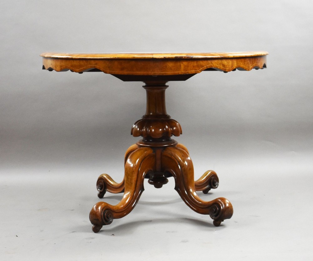 An oval Victorian burr walnut loo table featuring geometric and floral marquetry inlay, shown upright via a tilt-top mechanism on a carved four-legged pedestal base. View IV