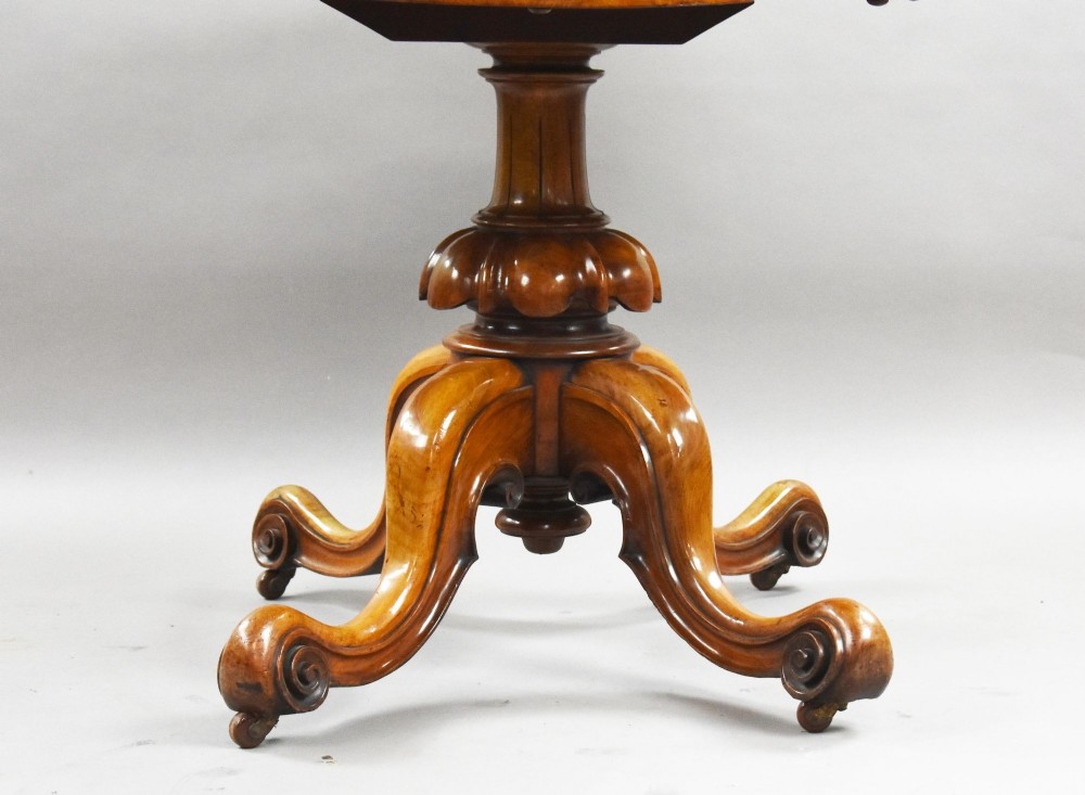 An oval Victorian burr walnut loo table featuring geometric and floral marquetry inlay, shown upright via a tilt-top mechanism on a carved four-legged pedestal base. View V