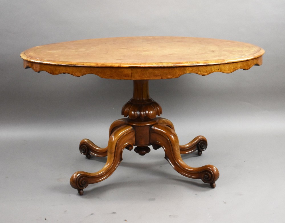 An oval Victorian burr walnut loo table featuring geometric and floral marquetry inlay, shown upright via a tilt-top mechanism on a carved four-legged pedestal base. View VI