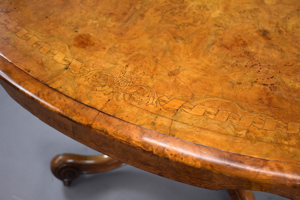 An oval Victorian burr walnut loo table featuring geometric and floral marquetry inlay, shown upright via a tilt-top mechanism on a carved four-legged pedestal base. View VII