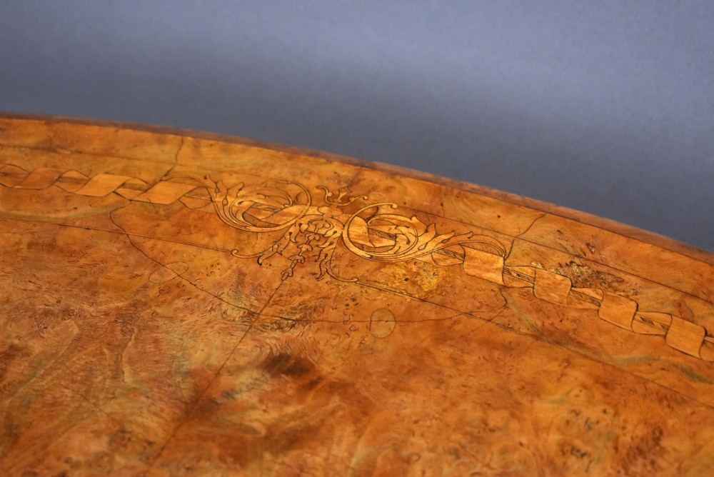 An oval Victorian burr walnut loo table featuring geometric and floral marquetry inlay, shown upright via a tilt-top mechanism on a carved four-legged pedestal base. View VIII