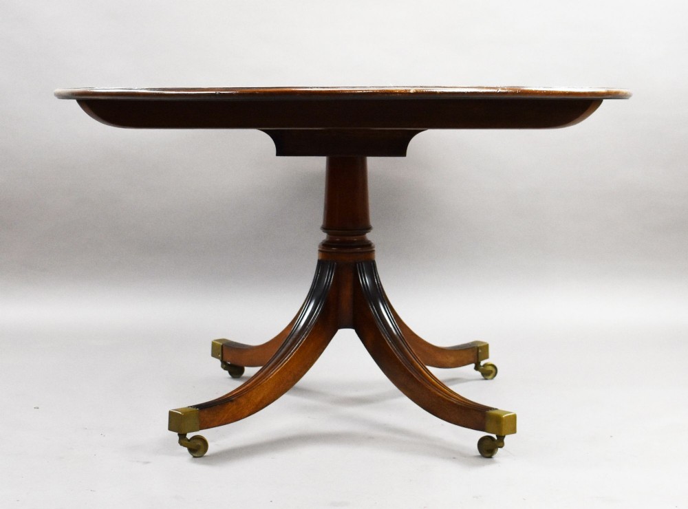 An oval Regency style flame mahogany tilt-top dining table featuring a crossbanded satinwood border and a pedestal base with brass castors. View III