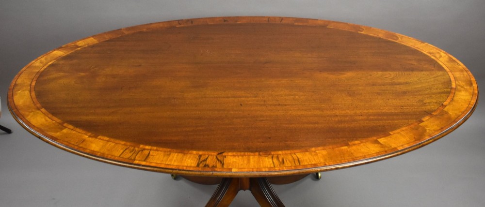 An oval Regency style flame mahogany tilt-top dining table featuring a crossbanded satinwood border and a pedestal base with brass castors. View VI