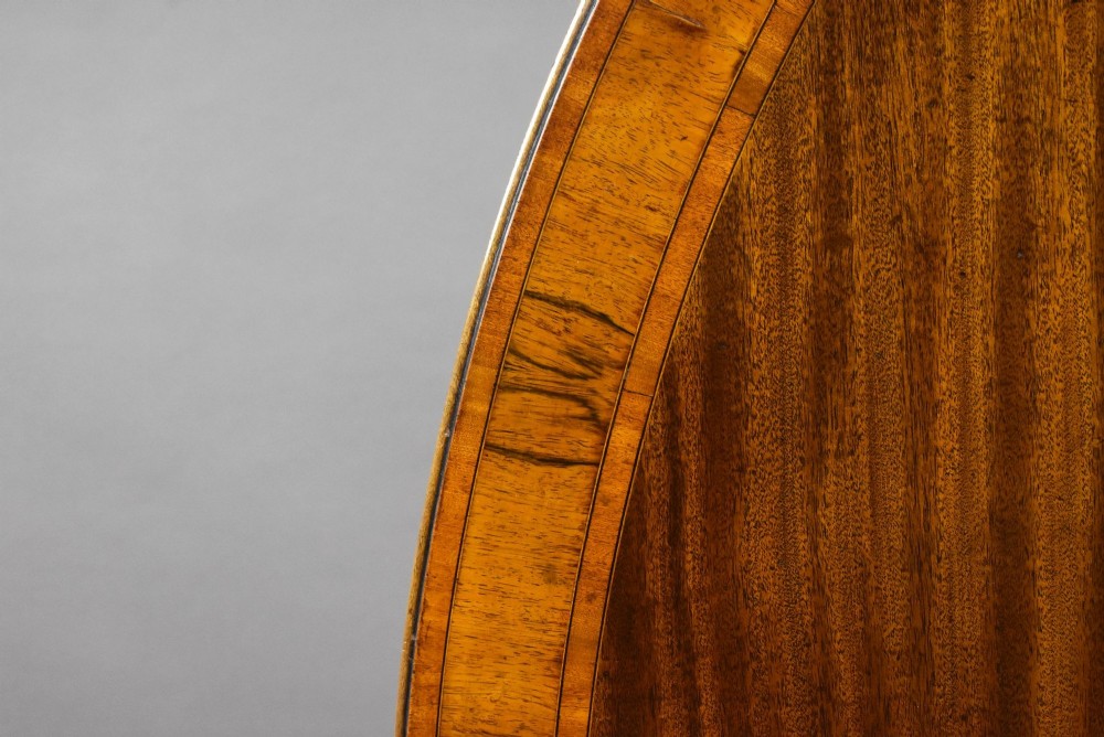 An oval Regency style flame mahogany tilt-top dining table featuring a crossbanded satinwood border and a pedestal base with brass castors. View VII