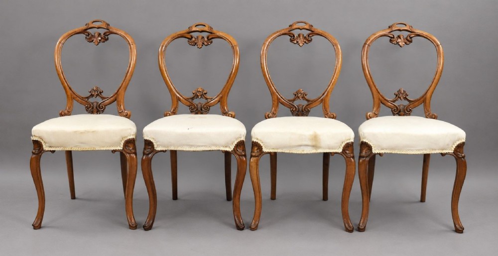 A set of four Victorian walnut balloon back dining chairs featuring carved back crests, cabriole front legs, and cream upholstered seats.