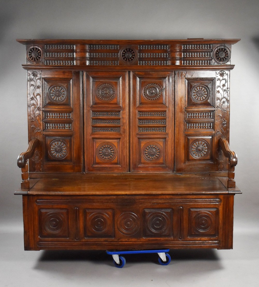 A high-backed dark oak panelled settle featuring geometric roundel carvings, a spindle frieze, scroll arms, and a box seat storage base. View II
