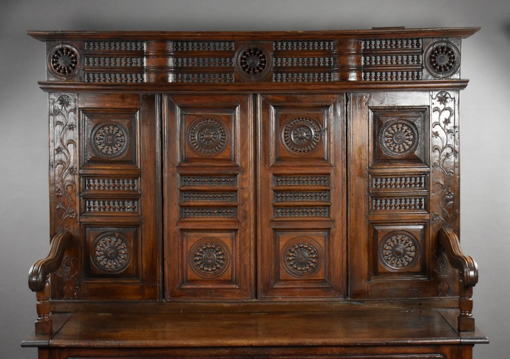 A high-backed dark oak panelled settle featuring geometric roundel carvings, a spindle frieze, scroll arms, and a box seat storage base. View III