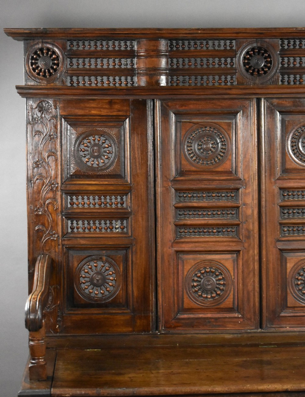 A high-backed dark oak panelled settle featuring geometric roundel carvings, a spindle frieze, scroll arms, and a box seat storage base. View IV